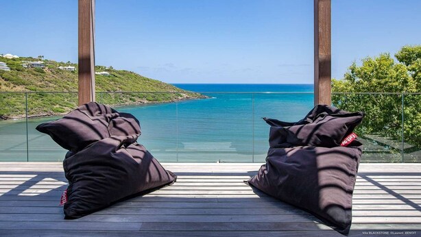 Ein Balkon mit zwei schwarzen Sitzsäcken, der Blick auf eine Küstenlandschaft mit klarem, blauem Wasser und hügeligem Gelände bietet.