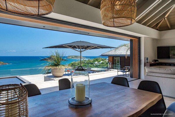 Un espace repas moderne avec vue sur l’océan, une grande plante en pot et un porte-bougie en verre sur la table.
