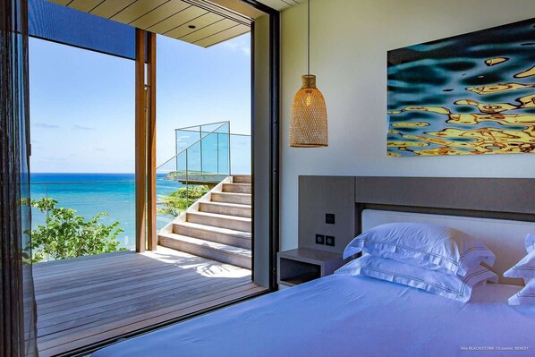 A modern bedroom with a large bed, a view of the ocean, a staircase leading to a glass structure, and a hanging light fixture.