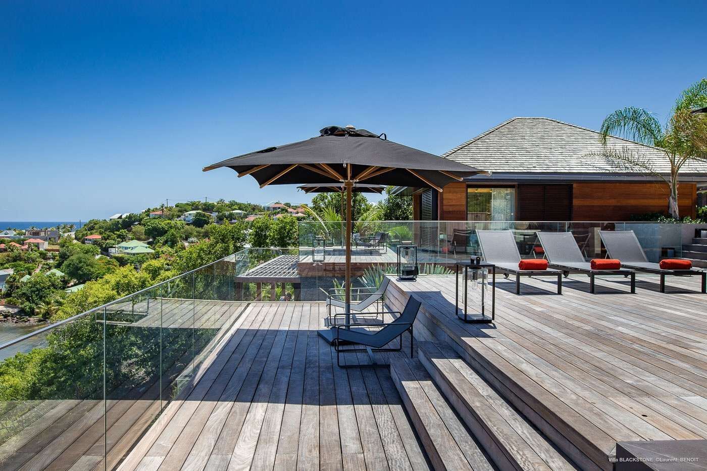Une terrasse en bois avec des chaises longues et un parasol, offrant une vue panoramique sur une charmante ville côtière.