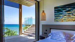 A modern bedroom with a large bed, a view of the ocean, a staircase leading to a glass structure, and a hanging light fixture. - Blackstone (4/6 Rooms) (Marigot)