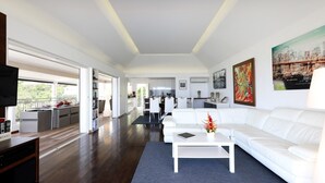 A spacious living area with a white sofa, a coffee table, and a TV. There is a kitchen in the background with white cabinets and a dining area. - Lenalee (2/4 Rooms) Luxury villa (Gustavia)