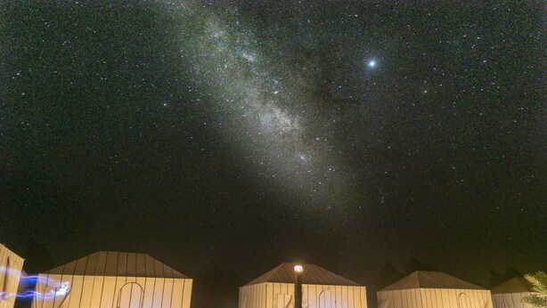 Céu noturno estrelado com a Via Láctea visível. Três grandes tendas com fontes de luz em suas bases.