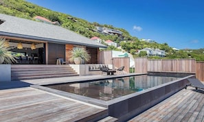 A modern outdoor swimming pool with wooden decking, a covered seating area, and a view of hillside residences.