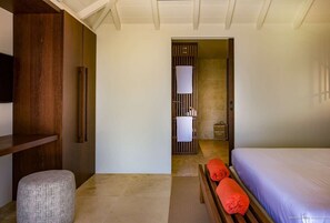 Een moderne hotelkamer met een houten bankje, een klein voetenbankje, een bed met een paars dekbed en een badkamer die door een open deur zichtbaar is.