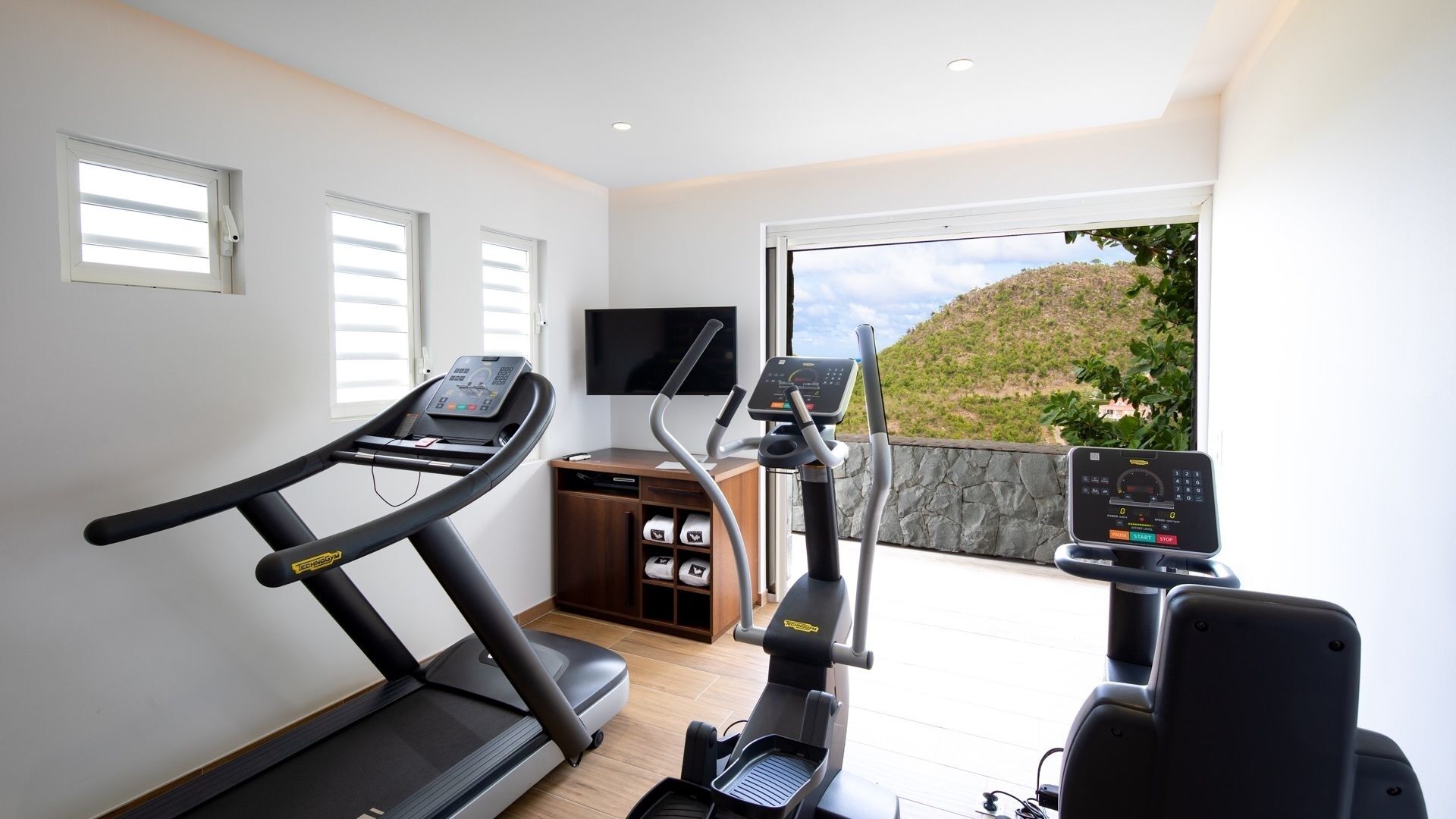 Een sportruimte met een loopband, elliptische trainer en hometrainer, een televisie en een stenen muur.