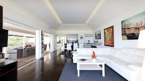 Un salón amplio con un sofá blanco, una mesa de centro y un televisor. Se ve una cocina al fondo con armarios blancos y una zona de comedor.
