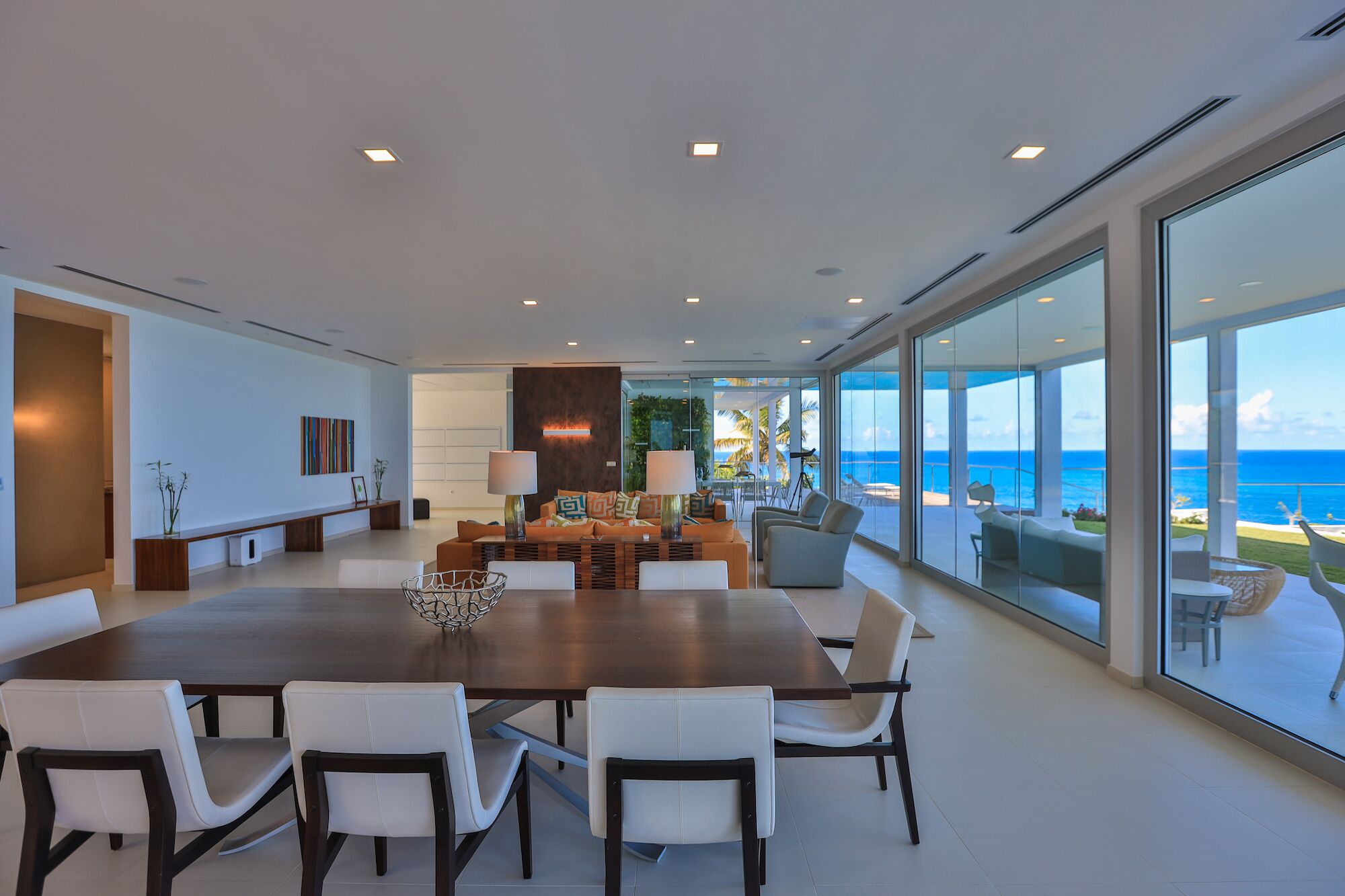 A modern dining area with a large table and chairs, a decorative centerpiece, and a view of the ocean through large windows.