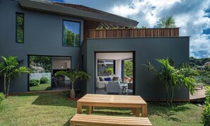 Een modern huis met een grote glazen deur die uitkomt op een open woonkamer, een houten terras en een kleine tuin met palmbomen.
