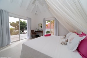 A bedroom with a large bed, a desk, and a chair. There is a view of palm trees and a building outside the window.