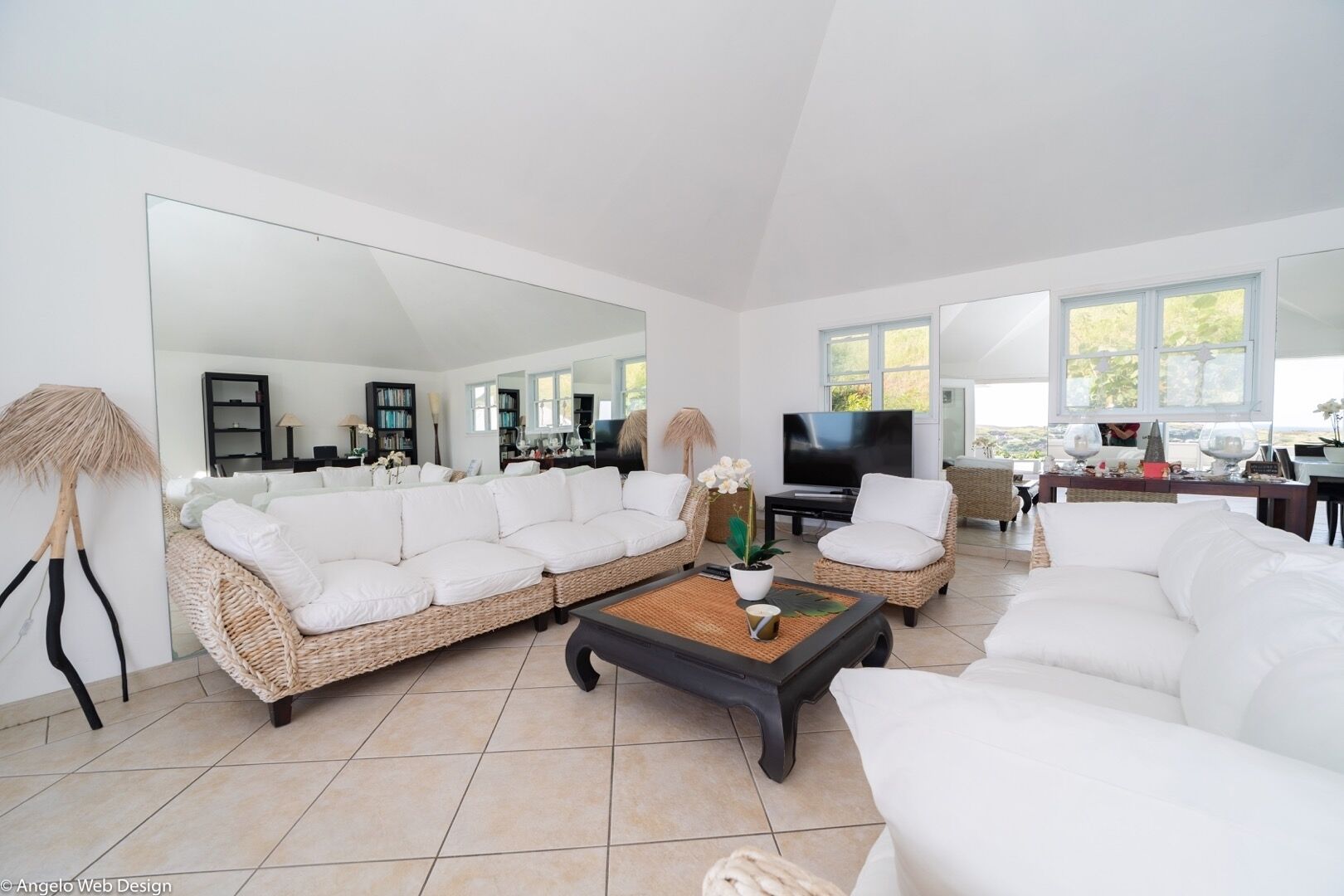 A spacious living room with a large sectional sofa, a coffee table, and a TV. There is a large mirror on one wall, and the room has a modern, minimalist design.