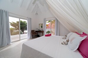 A bedroom with a large bed, a desk, and a chair. There is a view of palm trees and a building outside the window.