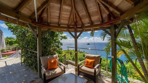 Área de estar coberta ao ar livre com duas cadeiras e uma mesa, com vista para a paisagem oceânica.