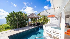 Piscina com um gazebo e espreguiçadeiras, cercada por vegetação e vista para o mar.