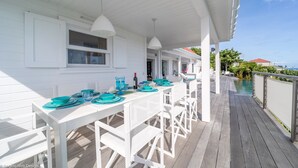 Un espace extérieur abrité, aménagé pour les repas, avec des tables et des chaises blanches, une piscine en arrière-plan et une vue sur l’océan.