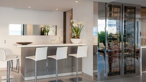 Eine moderne Küche mit weißer Arbeitsplatte, drei weißen Barhocker, einem großen Spiegel und einem Weinkühlschrank aus Glas.