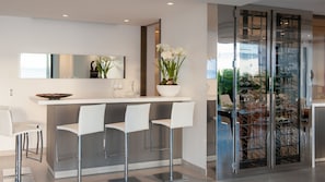 Eine moderne Küche mit weißer Arbeitsplatte, drei weißen Barhocker, einem großen Spiegel und einem Weinkühlschrank aus Glas.