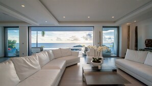 Ein modernes Wohnzimmer mit einer großen weißen Couch, einem Couchtisch und Meerblick durch raumhohe Fenster.