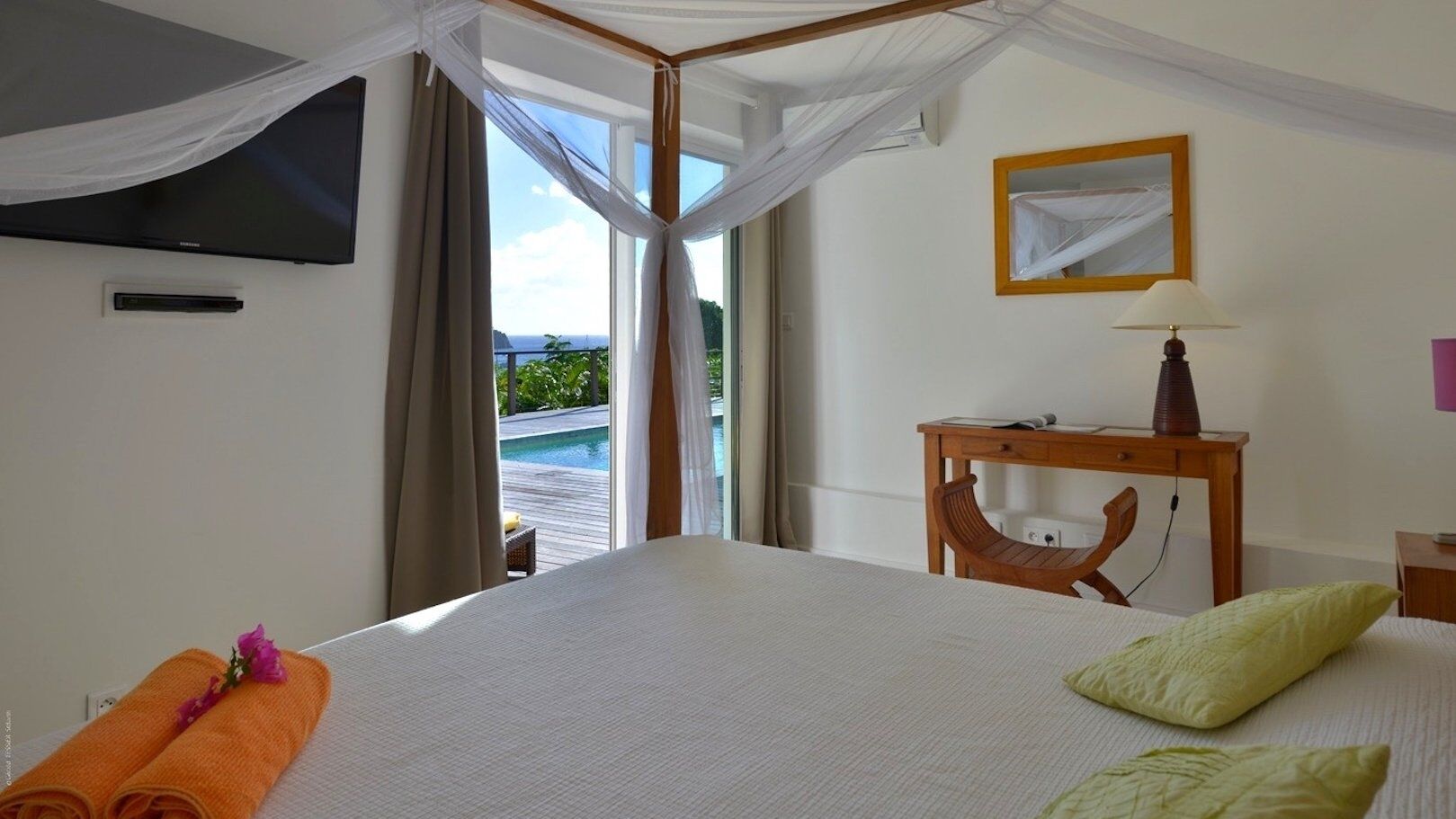 A hotel room with a bed, a wooden nightstand, a TV mounted on the wall, and a view of a pool through the window.