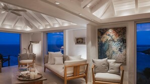 Un salon spacieux offrant une vue sur l’océan, avec un canapé en bois, un fauteuil et une table basse agrémentée de livres et de bougies.