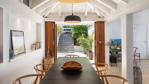 Een eethoek met een lange tafel, houten stoelen, een hanglamp en uitzicht op een trap die naar een balkon met planten leidt.