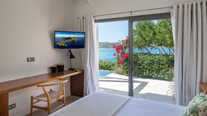 Een slaapkamer met een bed, een bureau, een stoel, een televisie en een schuifdeur die leidt naar een balkon met uitzicht op zee en groen.