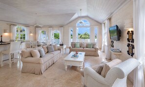A spacious living room with a high ceiling, white furniture, and a television mounted on the wall.