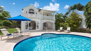 A pool area with a clear blue pool, white lounge chairs, and a white building with a balcony.