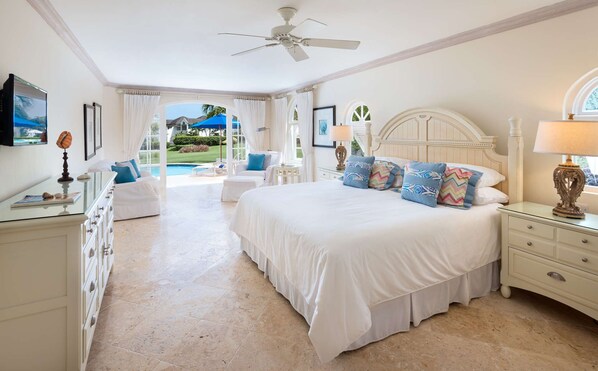 A spacious bedroom with a large bed, a ceiling fan, and a view of a pool and greenery outside.