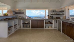 A spacious kitchen with a view of the sea, featuring white cabinetry, a central island, and various kitchen appliances. - Supersky Luxury villa (St barts)