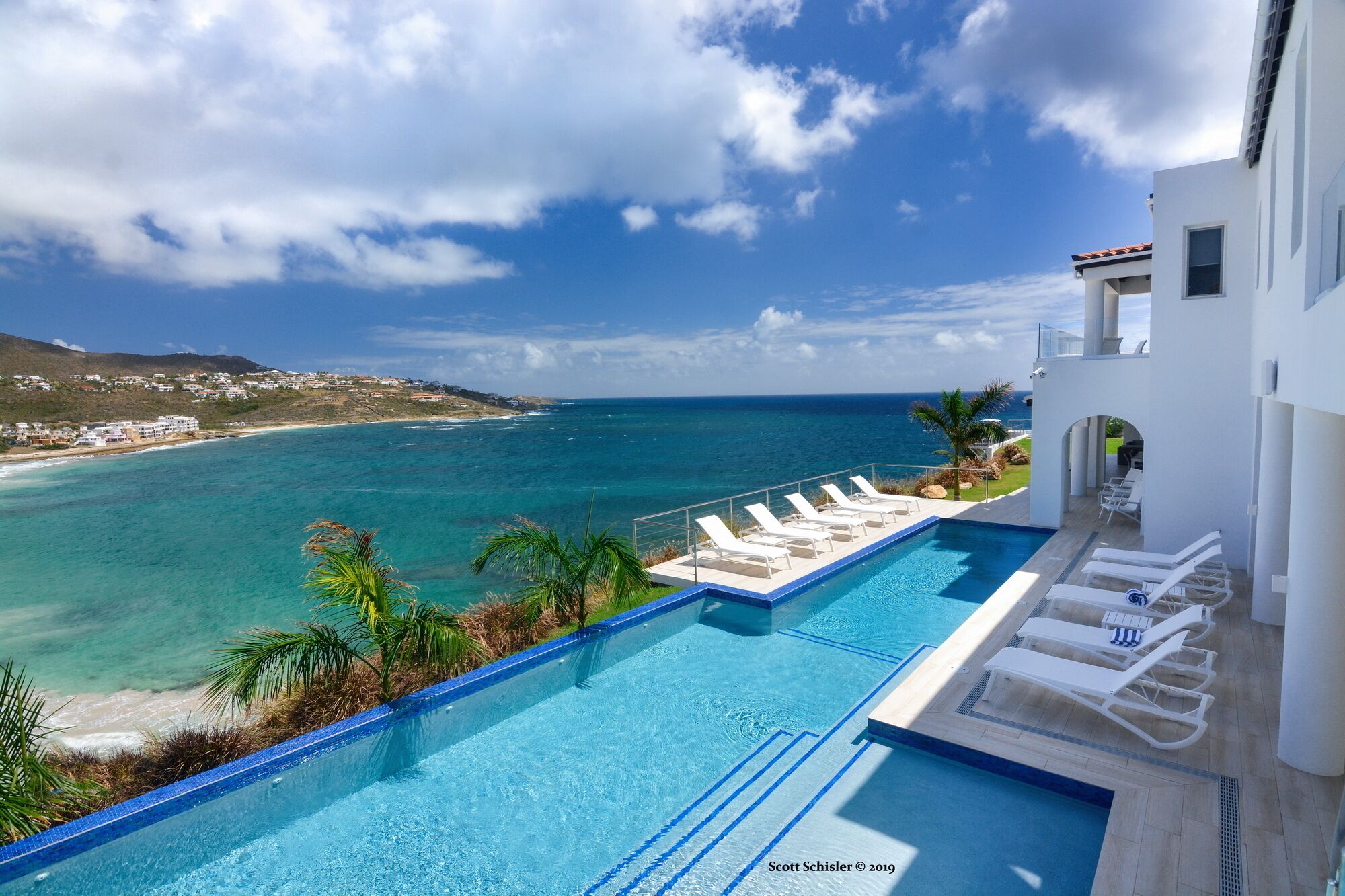 A pool with clear blue water, a view of the ocean, and a white building with a balcony.