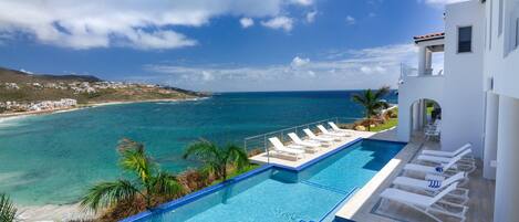 A pool with clear blue water, a view of the ocean, and a white building with a balcony.