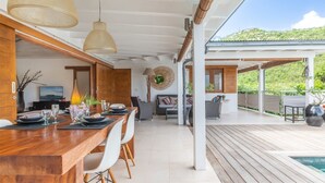 Een moderne eetruimte met een houten tafel, witte stoelen en hanglampen. Links is een zithoek met banken en een televisie, en rechts een houten terras met een zwembad.