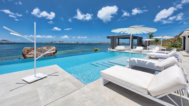 A modern infinity pool with white lounge chairs and umbrellas, overlooking a clear blue sea and a distant cityscape. - Mirabelle (3/5 Rooms) (Terres Basses)