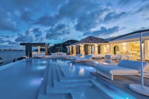 A luxurious outdoor pool area with modern lounge chairs and a dining area, overlooking a body of water and hills.