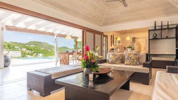 Amplia sala de estar con un gran ventanal con vistas a una piscina y follaje. Hay una mesa de centro de madera con un florero, un sofá con cojines estampados y un comedor con sillas.