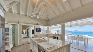 A modern kitchen with a large island, a dining area with outdoor seating, and a view of the sea. - Mirabelle (4/5 Rooms) (Terres Basses)
