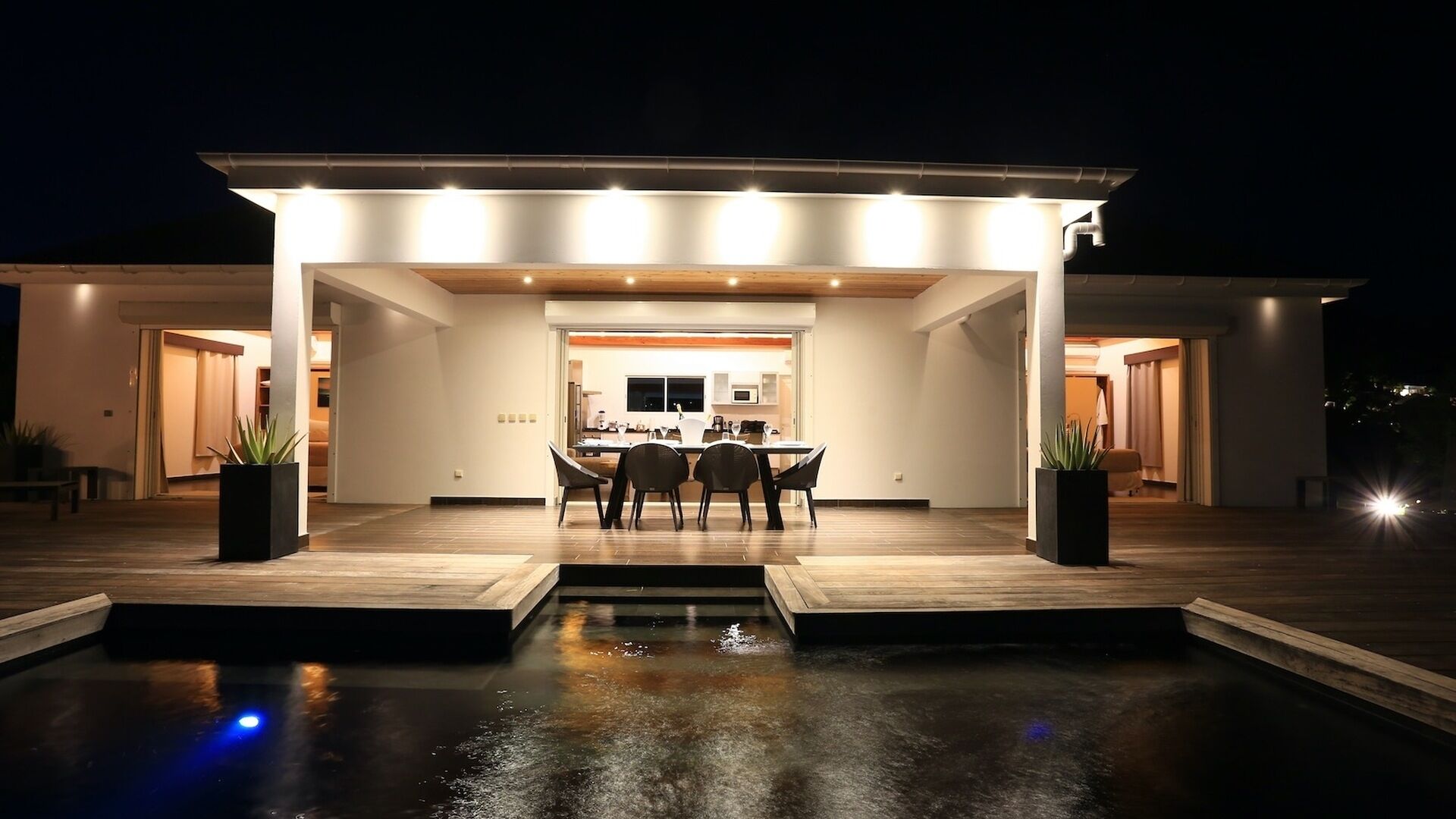 Une maison moderne dotée d’une piscine, d’un coin repas et d’une cuisine visible à travers une porte ouverte.