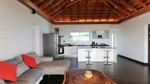 Eine moderne Küche mit einer zentralen Kochinsel und Barhocker, ein Essbereich mit einer Couch und roten Kissen sowie eine Holzbalkendecke.