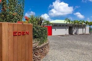 A wooden sign with "EDEN" in red letters, a gravel driveway, a building with a green roof, and palm trees. - Eden House (1/3 Rooms) (Marigot)