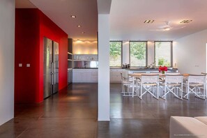 A modern kitchen with a red accent wall, a dining area with white chairs, and a large window with blinds. - Eden House (1/3 Rooms) (Marigot)