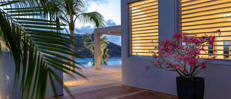 A modern villa with a wooden deck, large windows with horizontal blinds, and a view of a body of water and hills.