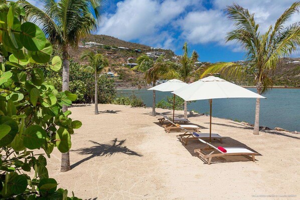Une plage avec des chaises longues en bois et des parasols blancs, un rivage sablonneux et une étendue d’eau en arrière-plan.