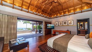 A spacious bedroom with a wooden ceiling, a large bed, a TV, and a view of the ocean through a sliding door. - Jade (4/8 Rooms) Luxury villa (Marigot)