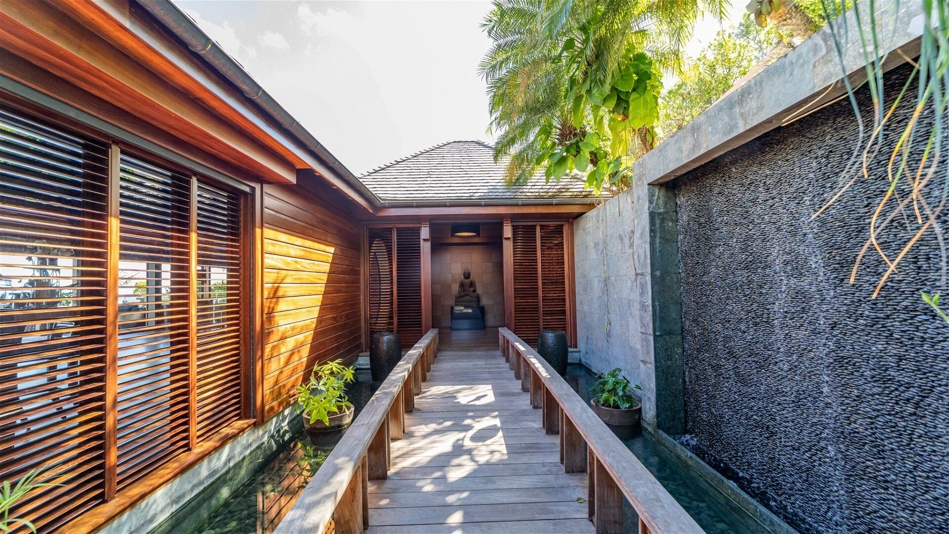 A wooden pathway leading to a meditation area with a statue, surrounded by wooden walls and a stone wall with a water feature.