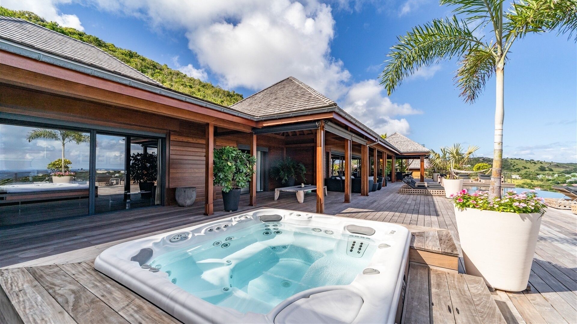 A modern outdoor hot tub area with a wooden deck, a covered seating area, and a view of the ocean.