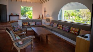 A spacious living area with a large sofa, wooden coffee table, and a chair. There is a window with a view of greenery outside. - Sucrier (1/2 Rooms) Luxury villa (Lorient)