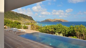 Une piscine à débordement avec une terrasse en bois, des chaises longues et une vue sur l’océan et les collines.