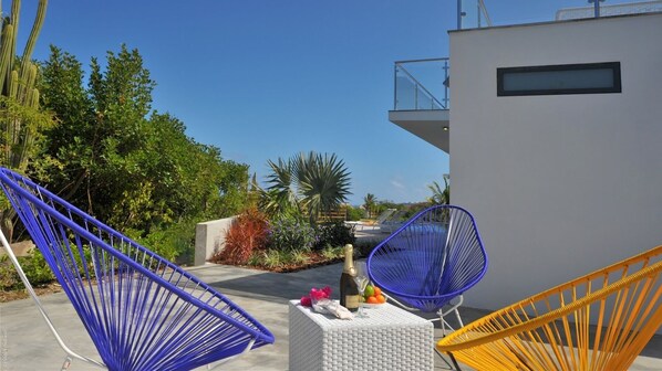 A modern outdoor seating area with blue and yellow chairs, a small table with a bottle and fruit, and a white building with a balcony.