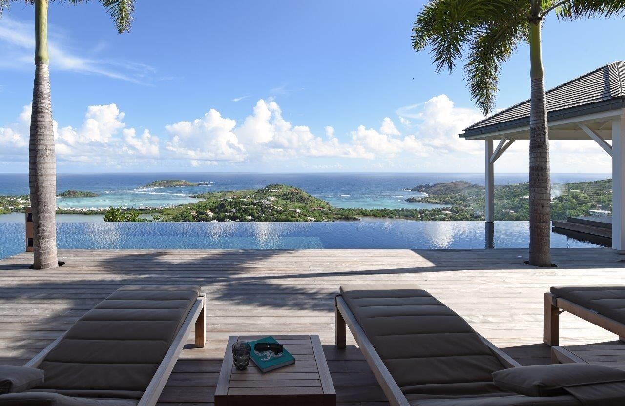 A wooden deck with lounge chairs and a table, overlooking a clear blue ocean and a tropical island.